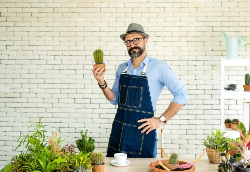 A man with a beard wearing a fedora and apron holding up a cactus in front of a collection of plants sitting on a table.