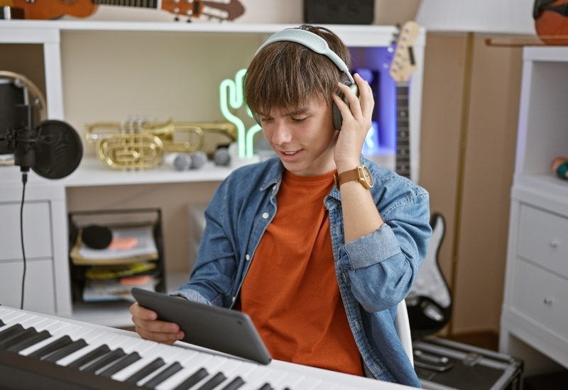 A teenager listening to music on his headphones while holding a tablet in a home studio surrounded by musical instruments.