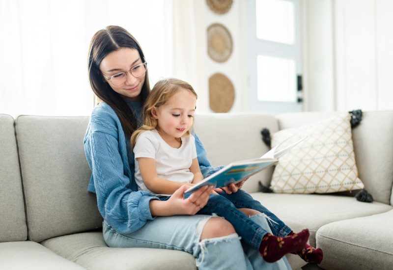 A teen babysitting her younger relative. The little girl is sitting on her lap, reading a book that the teen is holding open.