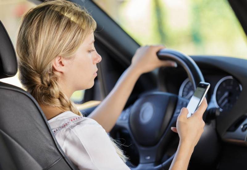 A teenage girl drives a car with one hand on the steering wheel, looking down at her smartphone in her right hand.